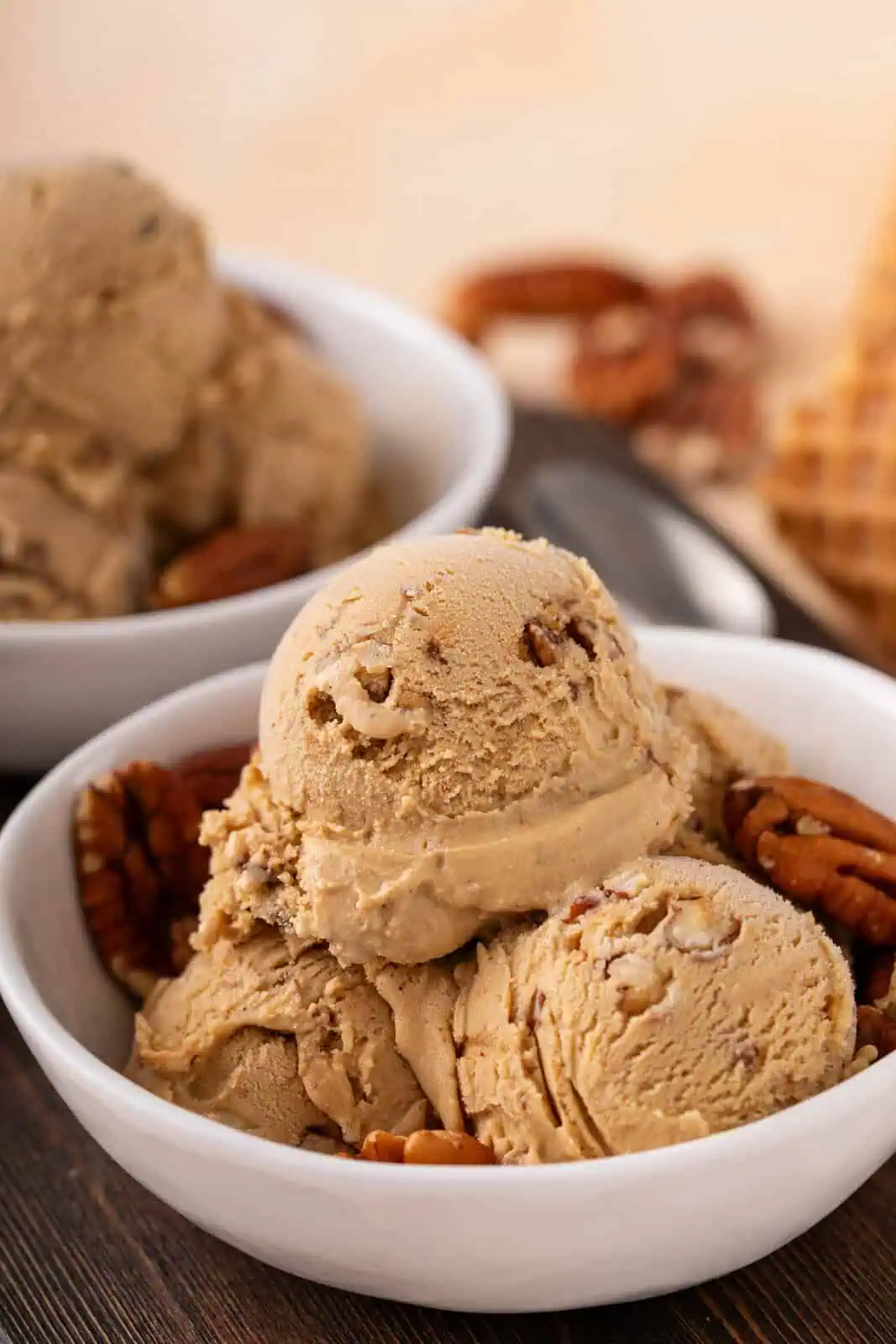 Side view of a white bowl with several scoops of butter pecan ice cream and pecan halves. A second bowl of ice cream is visible in the background.