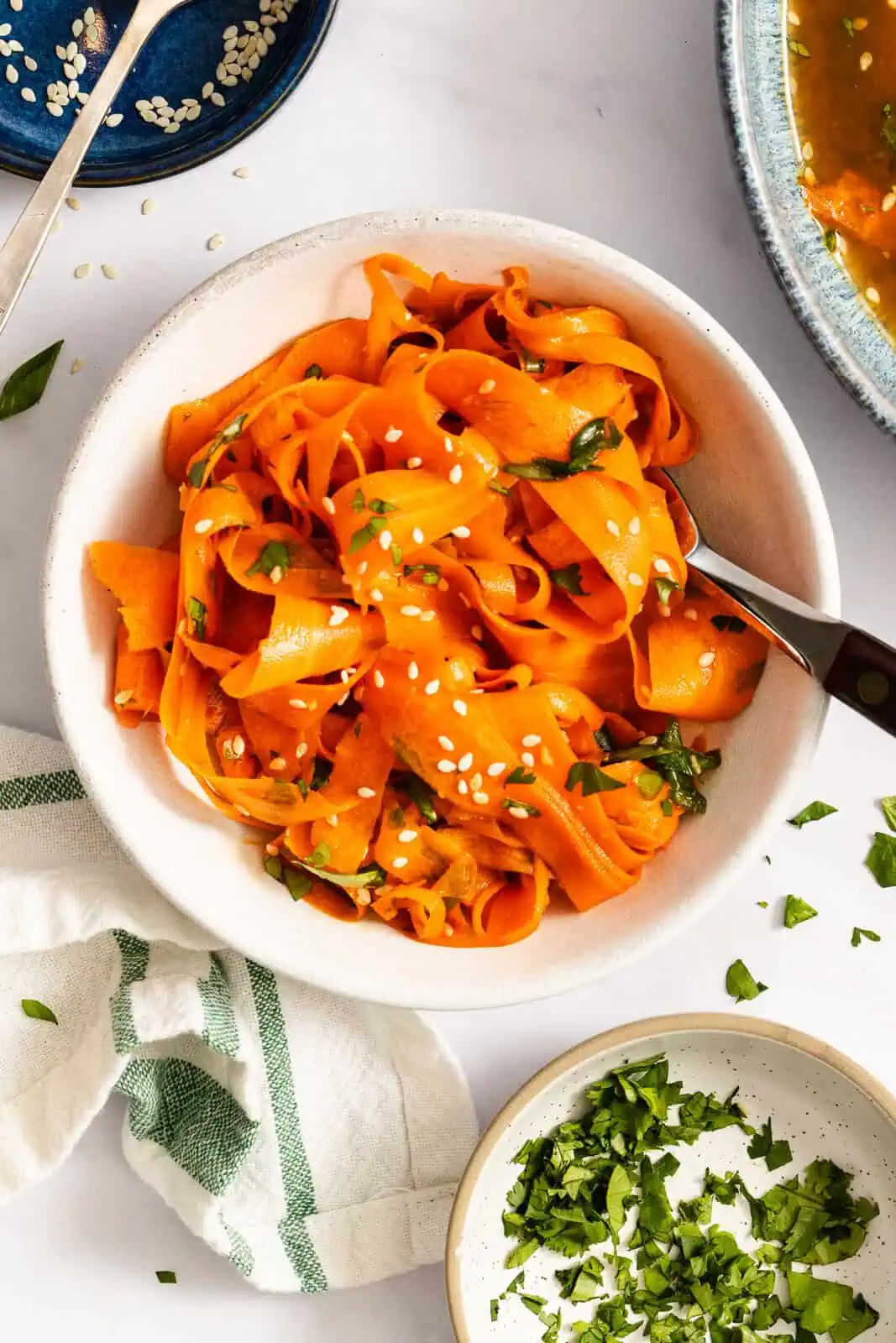 White bowl filled with shaved carrot salad, surrounded by bowls of fresh herbs.