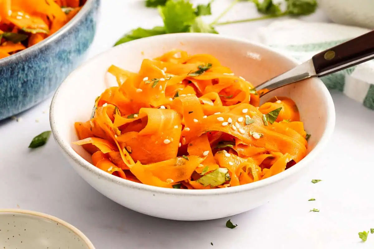 Side view of white bowl filled with shaved carrot salad, with a fork tucked into the bowl.