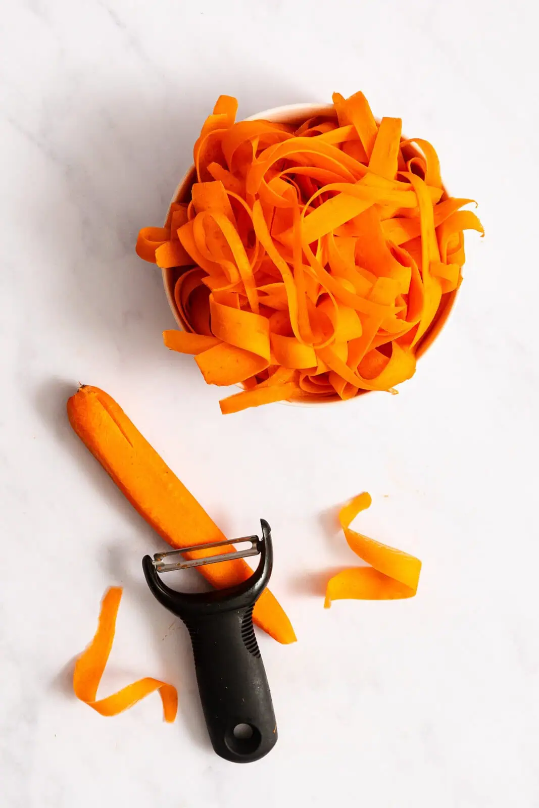 Bowl of shaved carrot ribbons next to a peeler showing how to peel carrot ribbons from a carrot on a countertop.