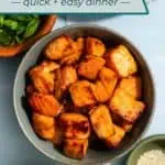 Bowl of air fryer salmon bites on a countertop surrounded by bowls of rice, scallions, cucumbers, and sesame seeds. Text overlay includes recipe name.