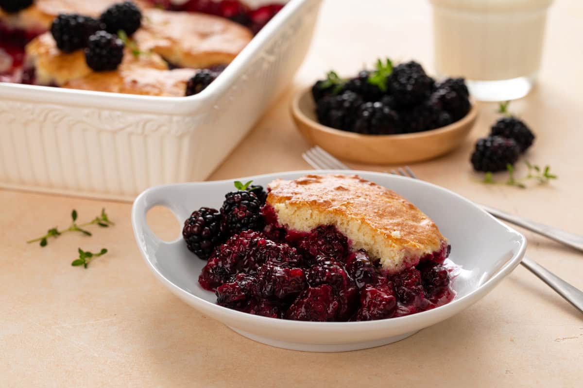 White bowl filled with a serving of blackberry cobbler, with a bowl of blackberries and the baking dish of the cobbler in the background.