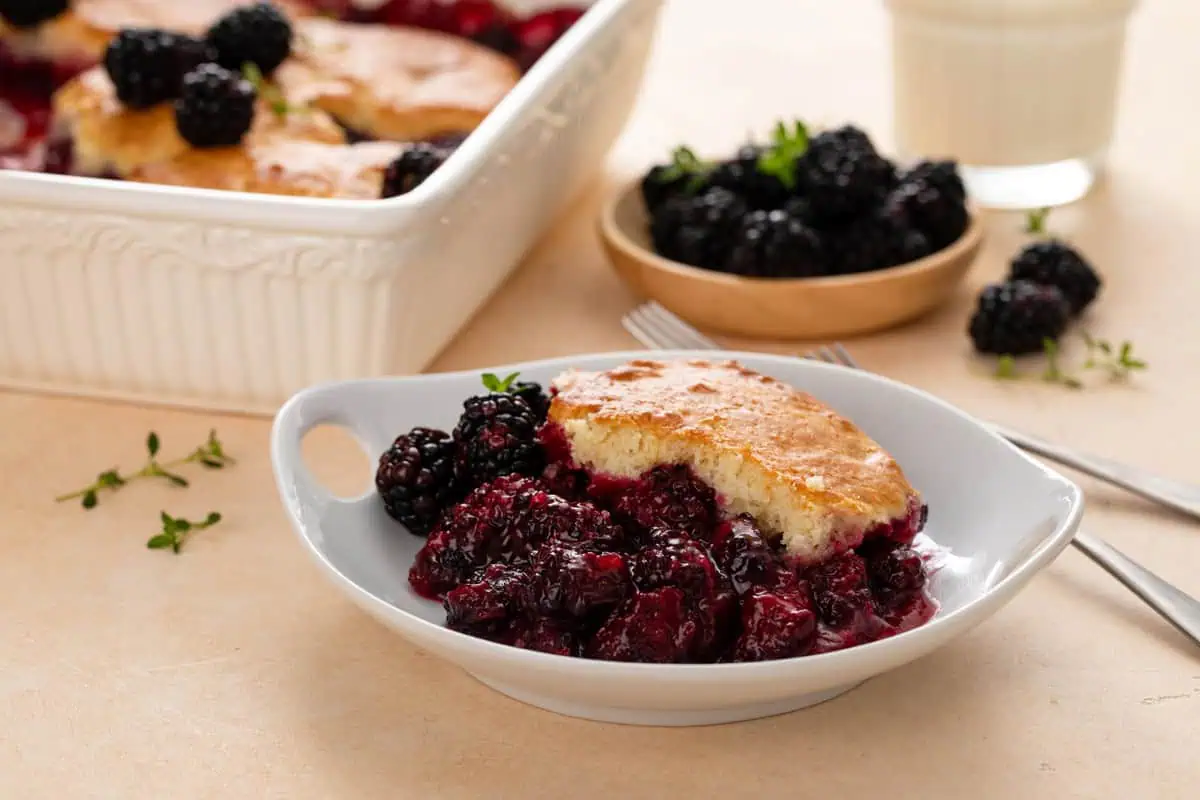 White bowl filled with a serving of blackberry cobbler, with a bowl of blackberries and the baking dish of the cobbler in the background.