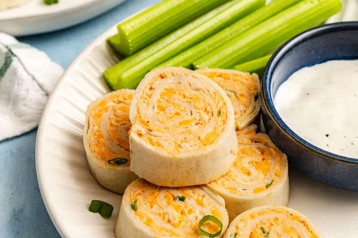 Close up of buffalo chicken pinwheel slices stacked on a white plate next to celery sticks.