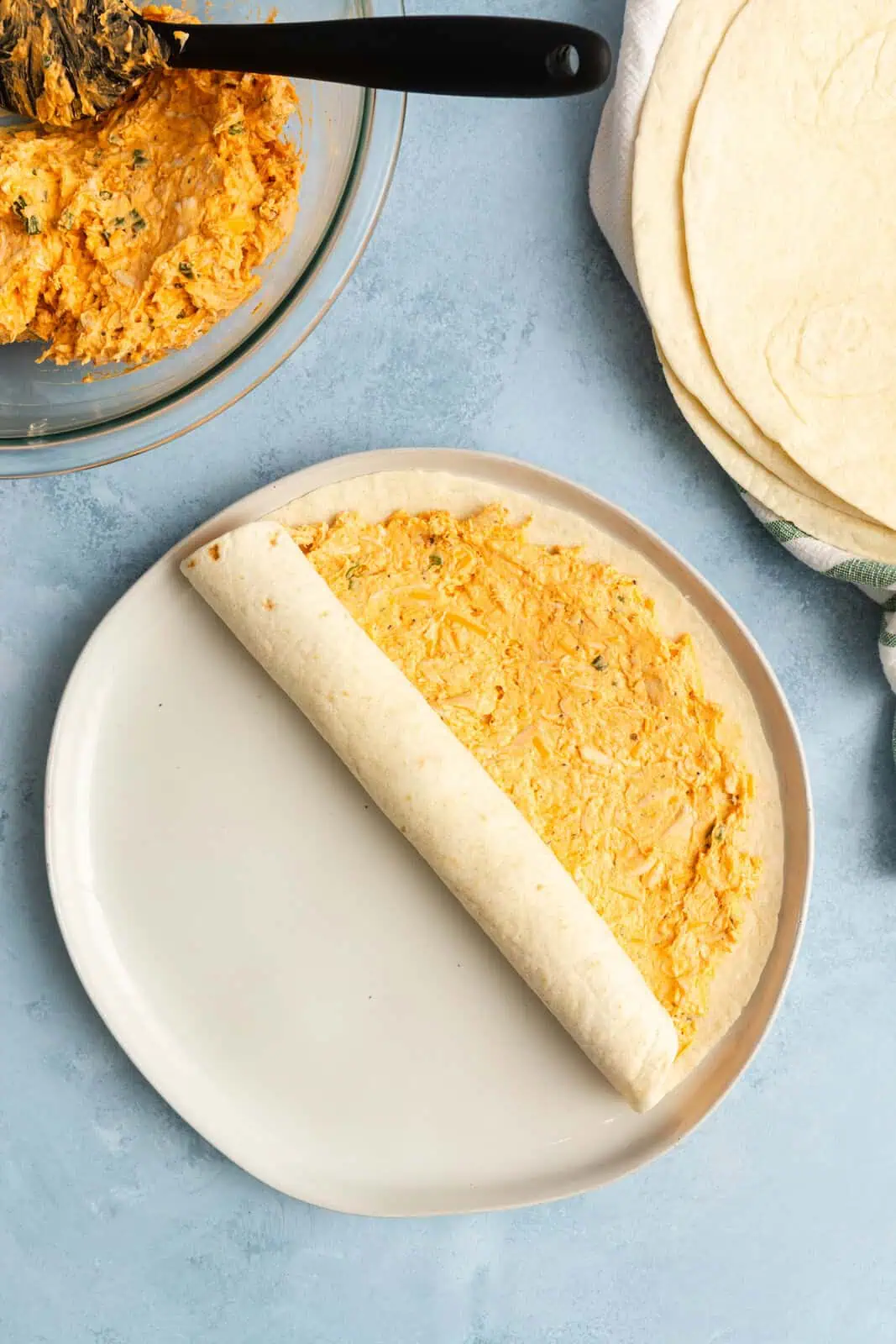 Tortilla being rolled up around buffalo chicken filling on a white plate.