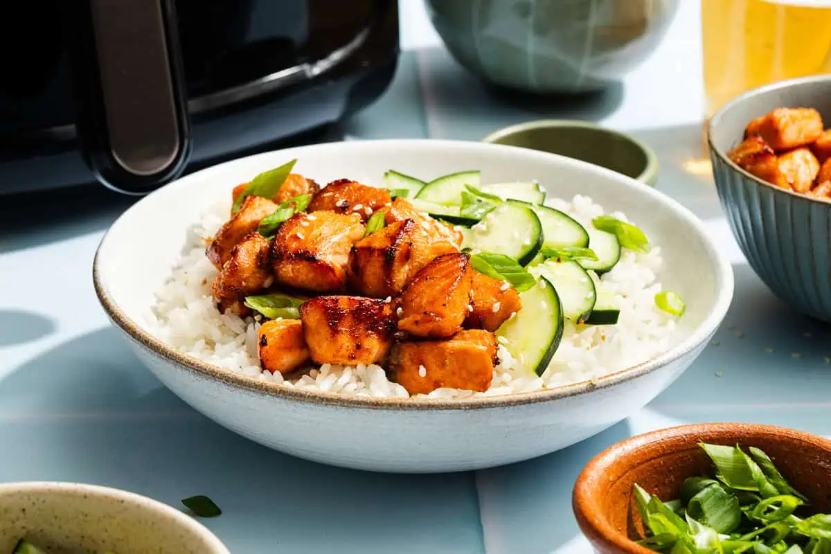 Salmon bites on a bed of rice and cucumbers in a white bowl, with the basket of an air fryer in the background.