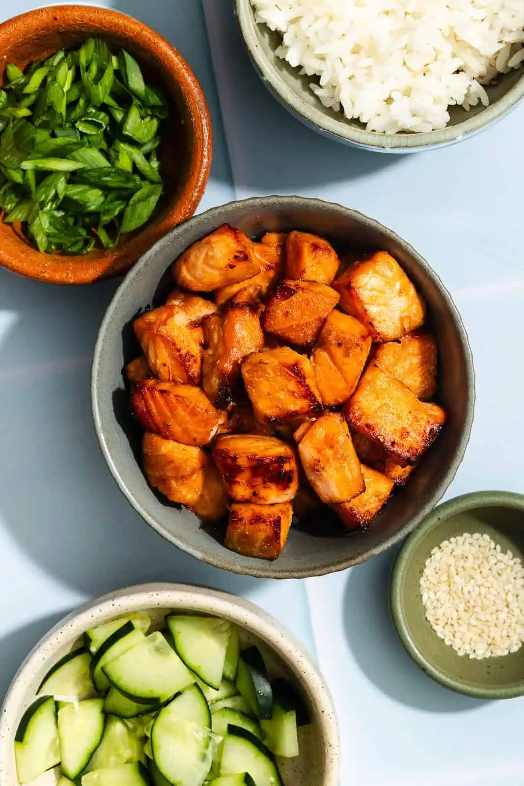 Bowl of air fryer salmon bites on a countertop surrounded by bowls of rice, scallions, cucumbers, and sesame seeds.