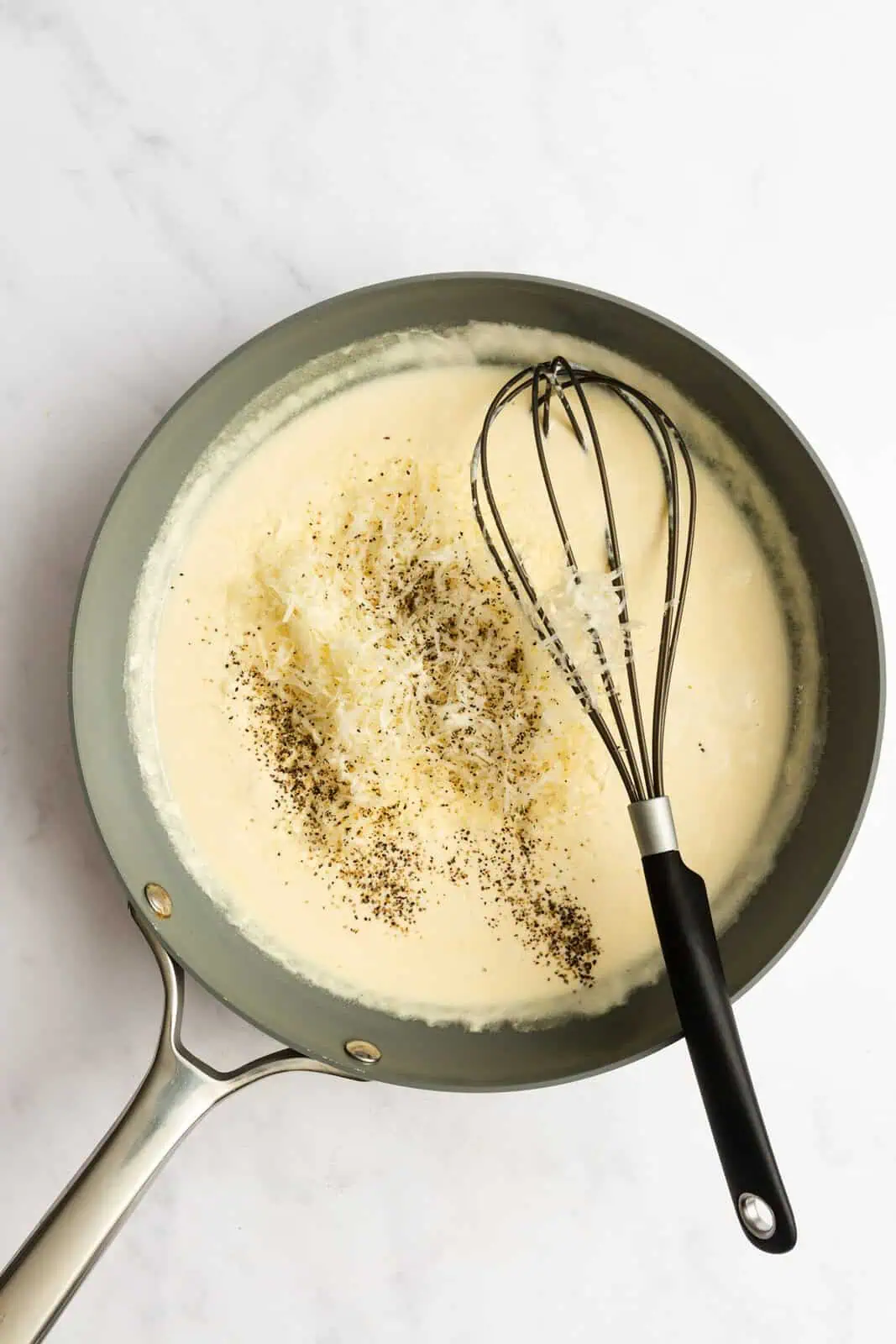 Salt and pepper being whisked into alfredo sauce in a skillet.