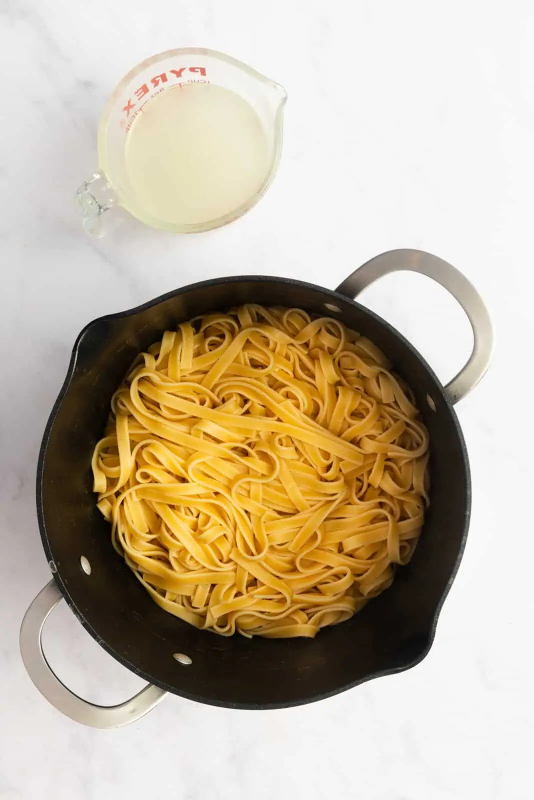 Cooked fettuccine pasta in a pot with a measuring cup of the cooking water sitting next to the pot.