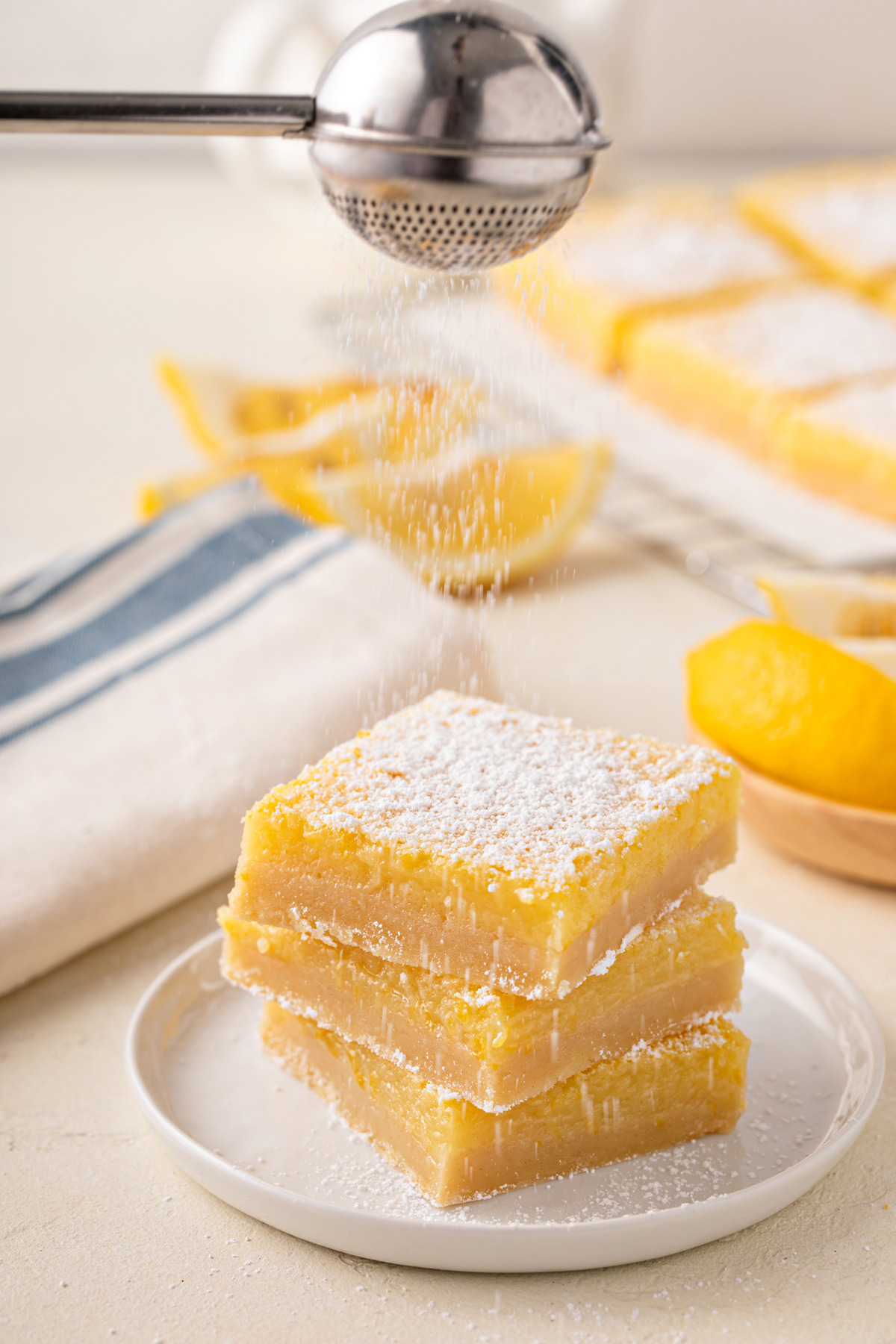Powdered sugar being dusted over three stacked lemon bars on a white plate.