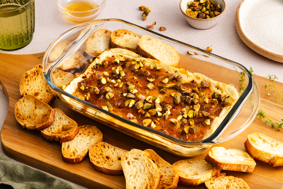 Baking dish of baked goat cheese dip on a wooden board, surrounded by slices of toasted baguette.