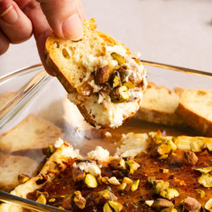 Hand holding a slice of crostini topped with baked goat cheese dip garnished with chopped pistachios.