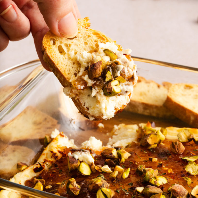 Hand holding a slice of crostini topped with baked goat cheese dip garnished with chopped pistachios.