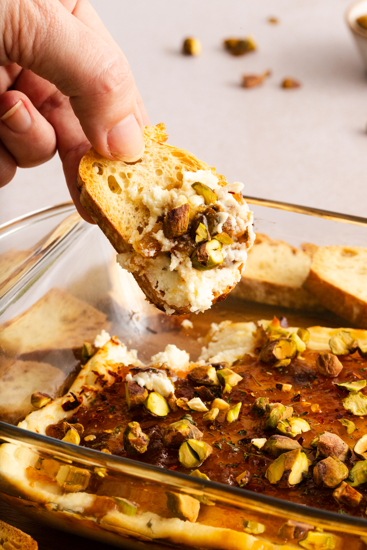 Hand holding a slice of toasted baguette scooped with baked goat cheese dip over a baking dish of the dip.