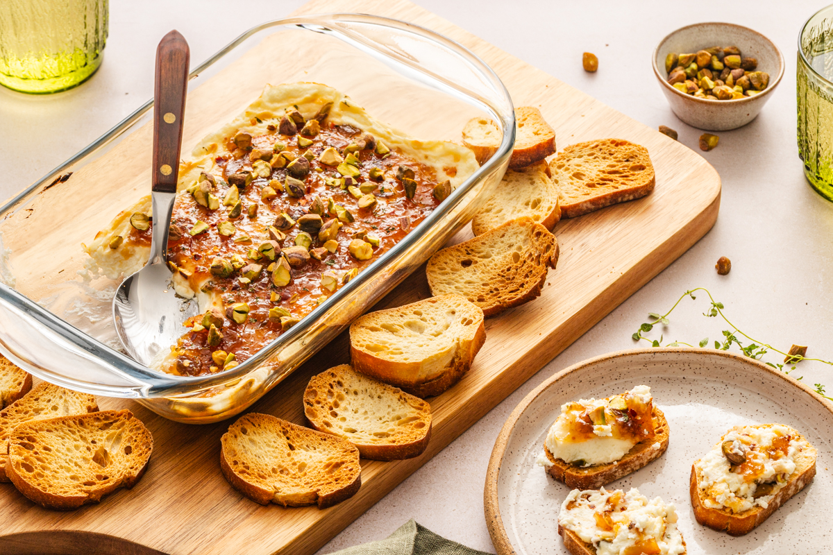 Wooden board holding a baking dish of baked goat cheese dip and crostini set next to a plate of crostini topped with the dip on a countertop.
