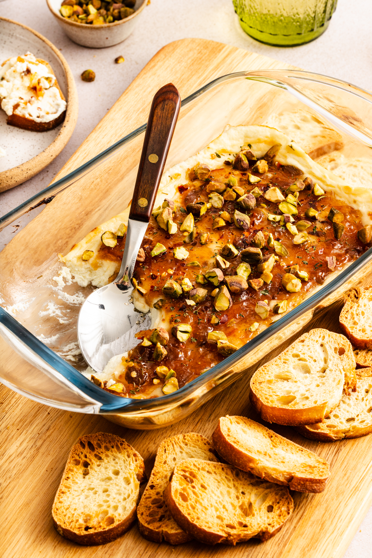 Spoon in a baking dish of baked goat cheese dip, surrounded by toasted baguette slices.